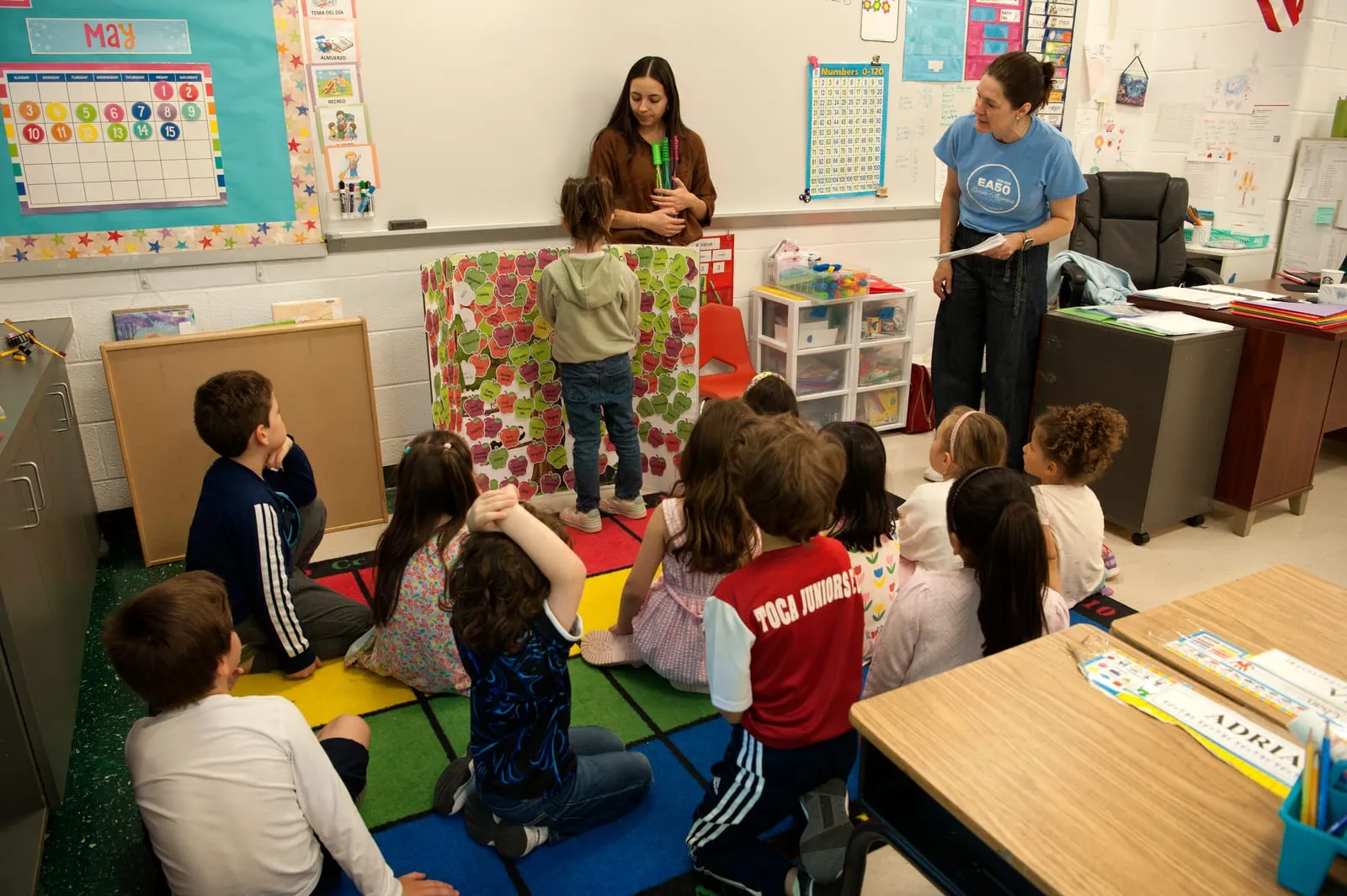 Una maestra presenta una actividad a un grupo de alumnos sentados en el piso del aula.