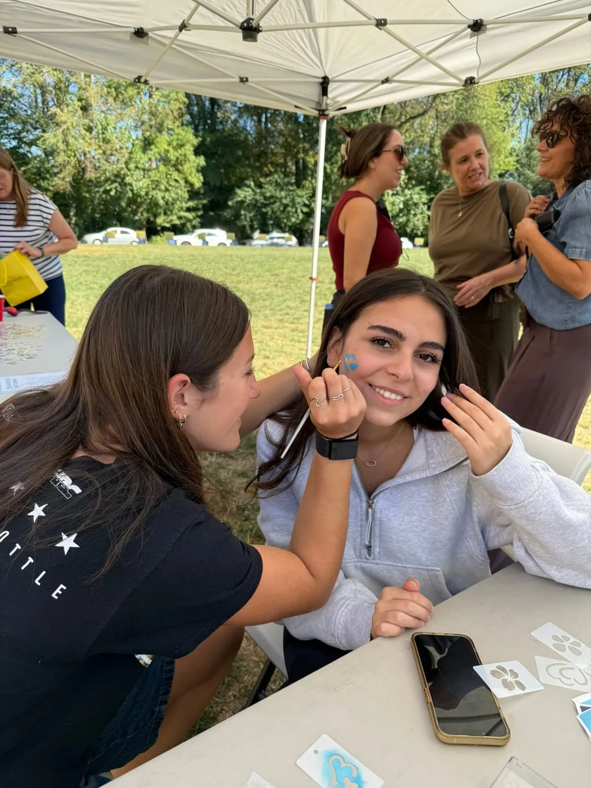 Una alumna pinta la cara de una compañera en un puesto bajo una carpa, durante el picnic del 25 de Mayo.